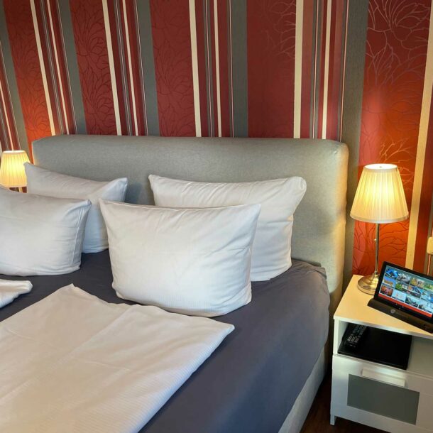 Doppelbett in der Junior Suite im Vitalhotel Schulenberg mit einer roten blumigen Tapete und auf jeder Bettseite ein kleiner Tisch mit einer Lampe