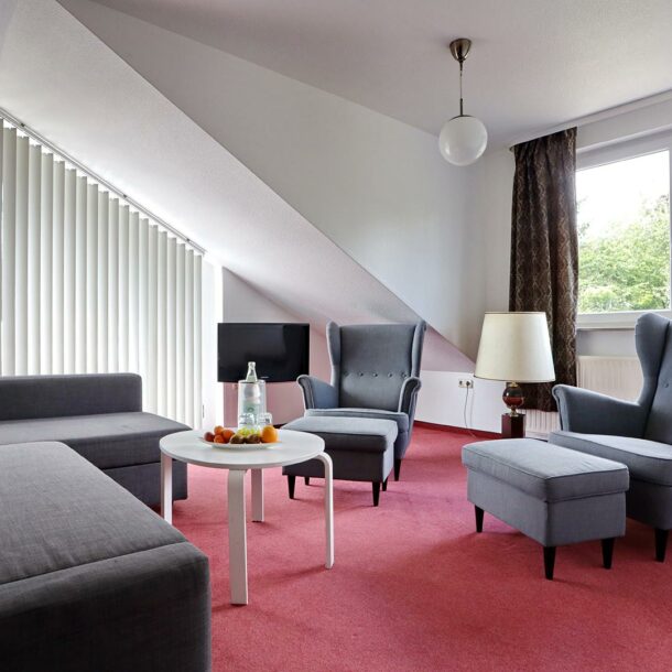 Zimmer der Suite Premium Plus im Vitalhotel Schulenberg im Harz mit einer grauen Couchgarnitur auf rotem Teppich. Zusätzlich ein weißer kleiner Couchtisch mit einem Obstkorb und einer Wasserflasche, dahinter an der Wand montiert ein Fernseher.