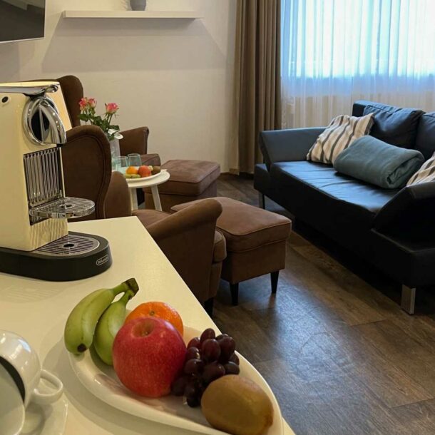 Der Wohnraum in der Junior Suite Premium mit einer Dunkelblauen und braunen Couchgarnitur, die mit der Couchlehne an dem weißen Bett steht. Der Raum hat dunkles Laminat und weiße Wände, sowie eine kleine Kaffeeecke mit Obst und einer Kaffeemaschine