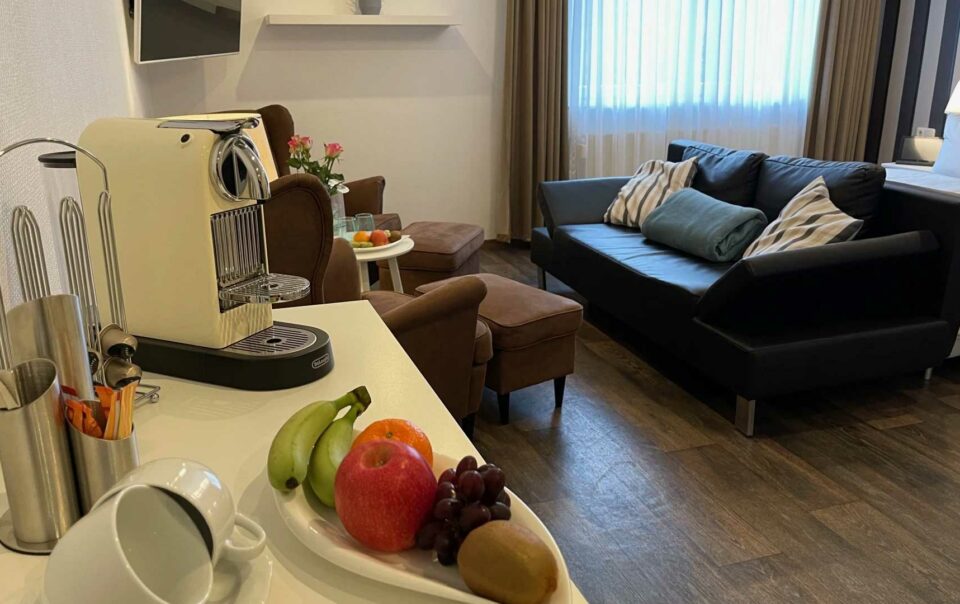 Der Wohnraum in der Junior Suite Premium mit einer Dunkelblauen und braunen Couchgarnitur, die mit der Couchlehne an dem weißen Bett steht. Der Raum hat dunkles Laminat und weiße Wände, sowie eine kleine Kaffeeecke mit Obst und einer Kaffeemaschine